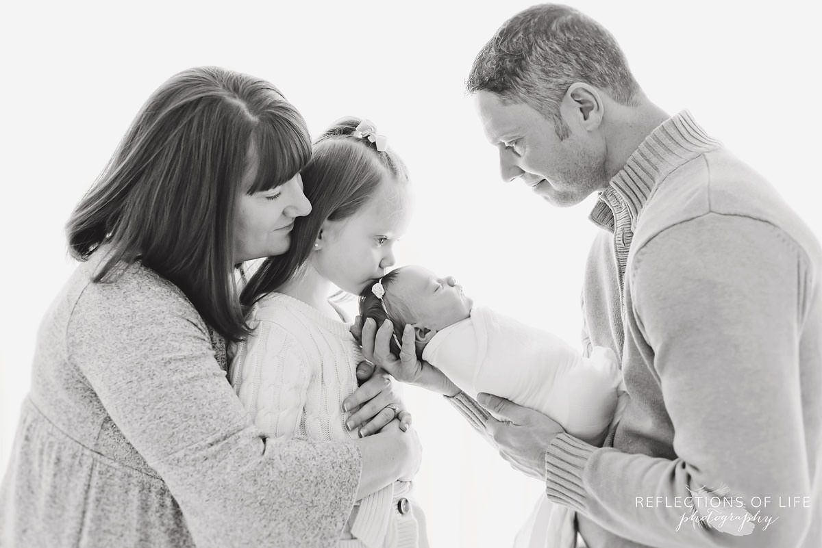 Family of four observes baby while daughter kisses their head black and white