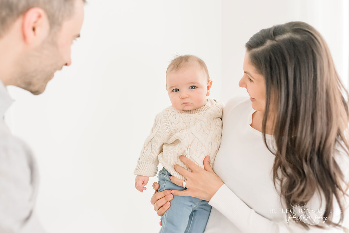mother holds baby as father smiles at him in natural light studio