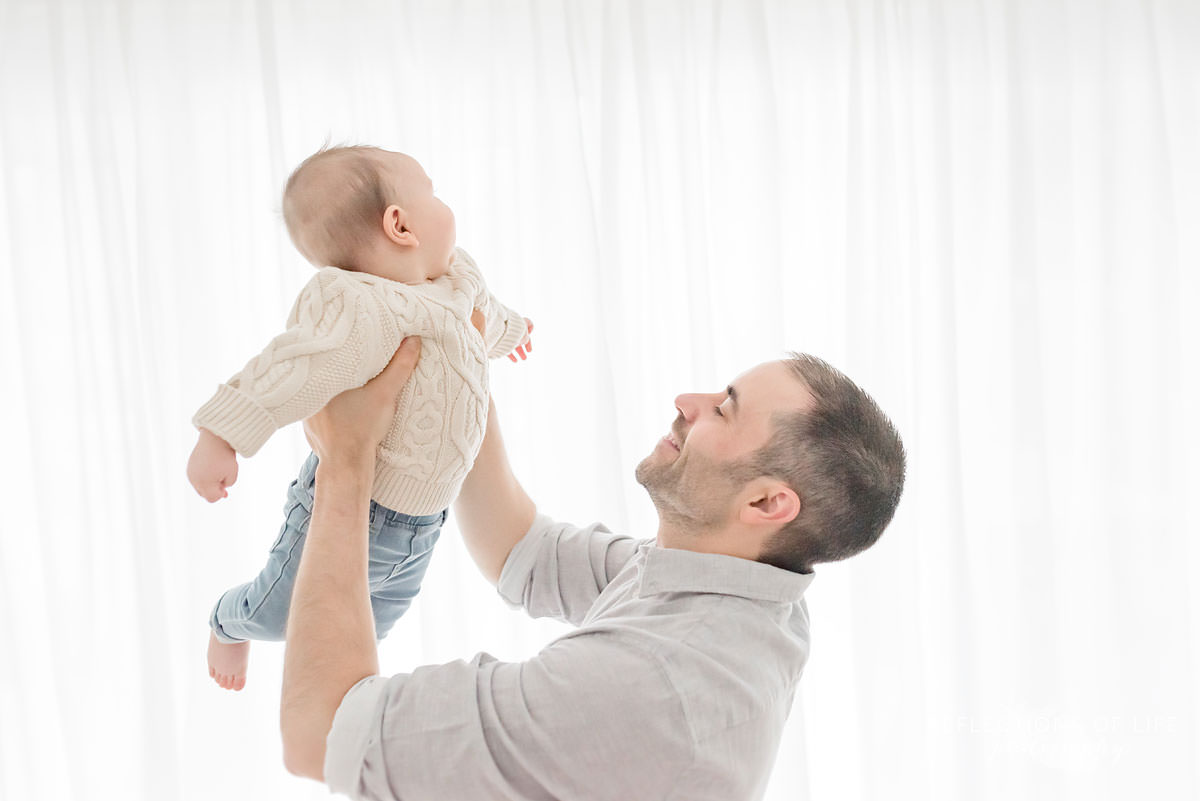 father lifts baby up as baby laughs in natural light studio