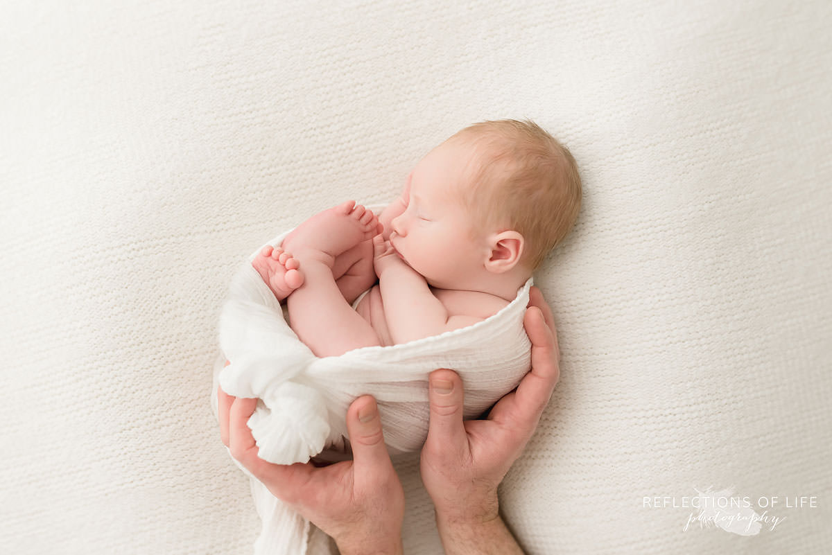 baby sleeps curling up in blanket with dad's hands surrounding him