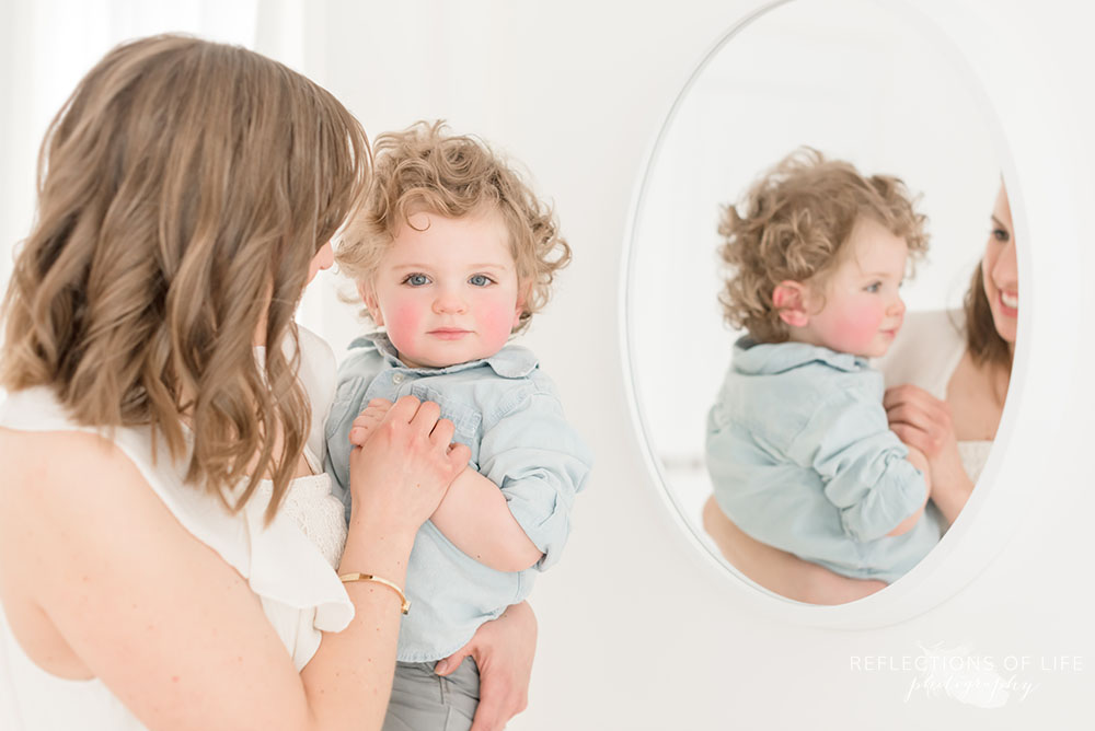 mother and son pose for the camera with a mirror reflection of cute little boy