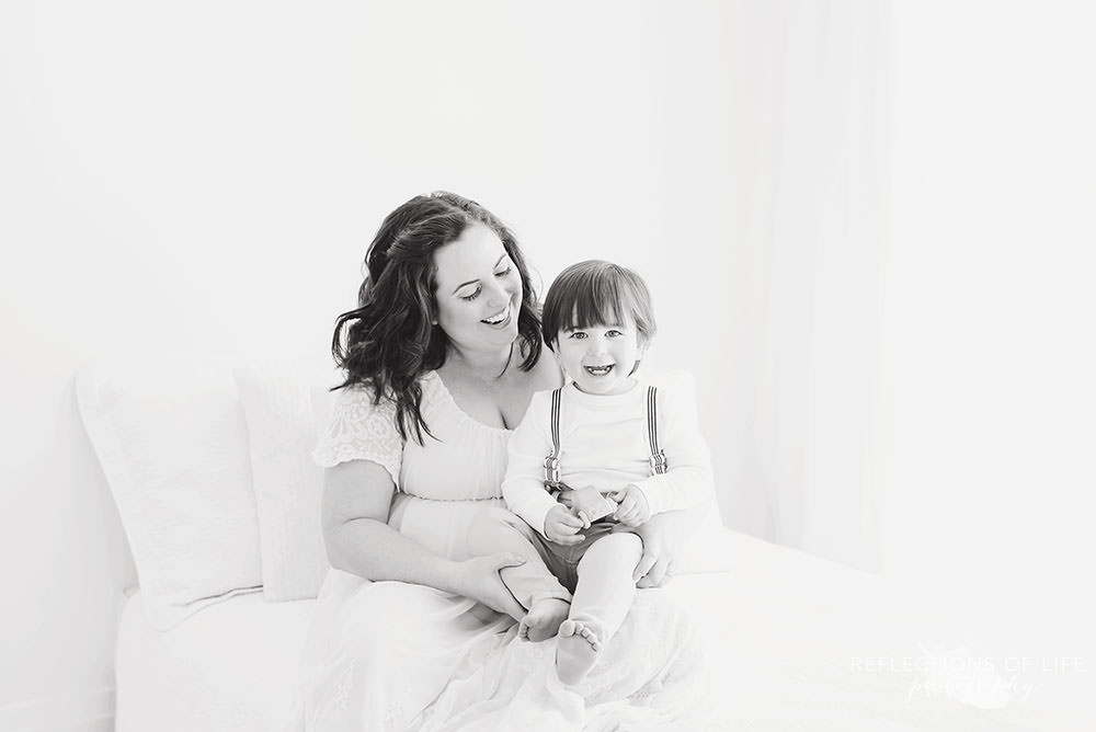 little boy smiles at the camera while mama smiles at him in natural light studio