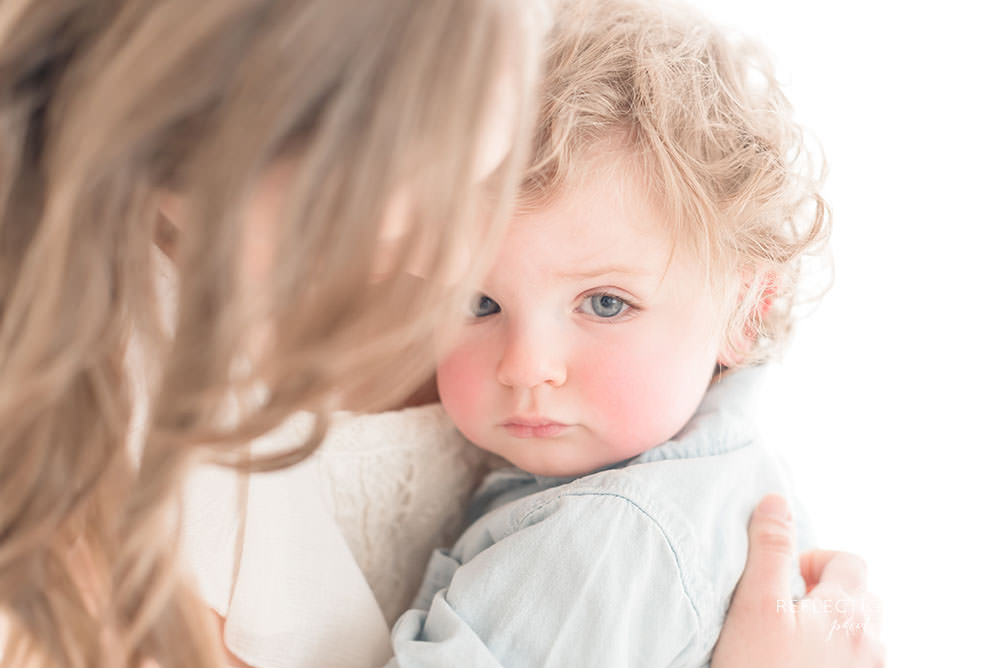 adorable little boy gazes into the camera with his mother holding onto him