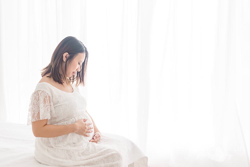 mama looks and feels her bump in natural light studio while her sits on the bed