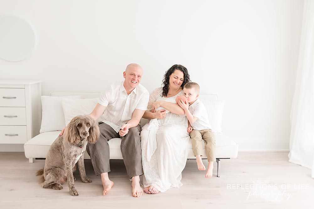 family of four sit on a couch as dad pets their dog in natural light studio