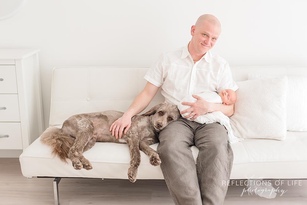 dad and baby sit on the couch with their family dog laying on his lap