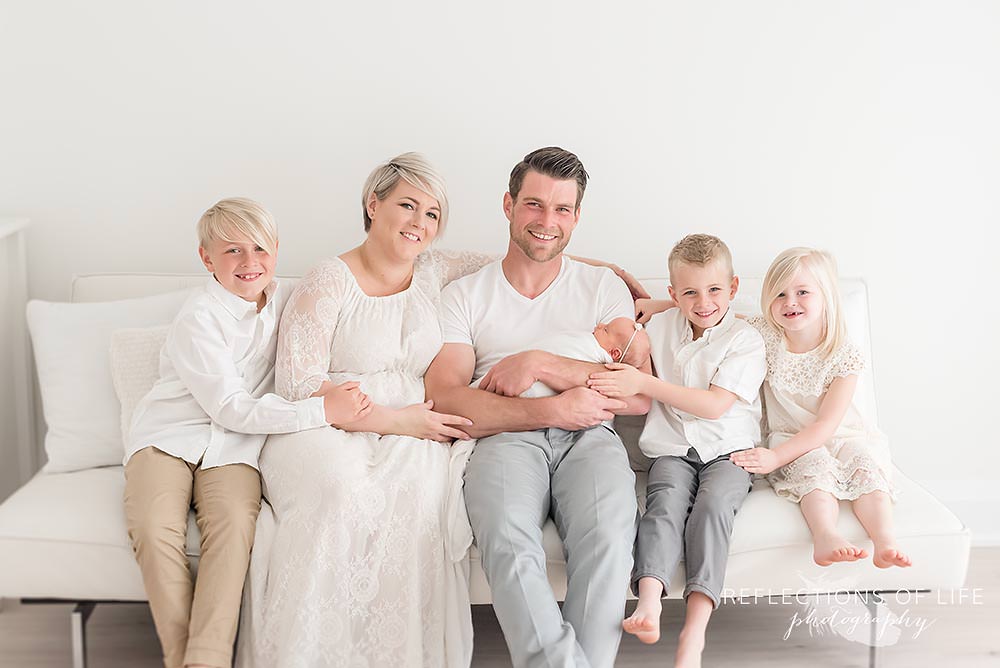 family of six sits on a couch holding their new daughter in natural light studio