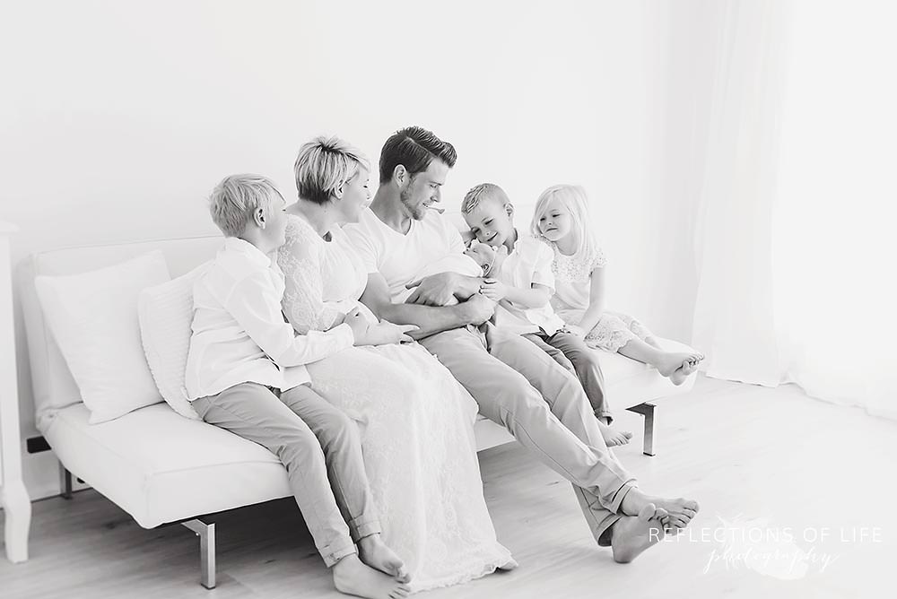 family of six sits on a couch looking at their newborn in black and white