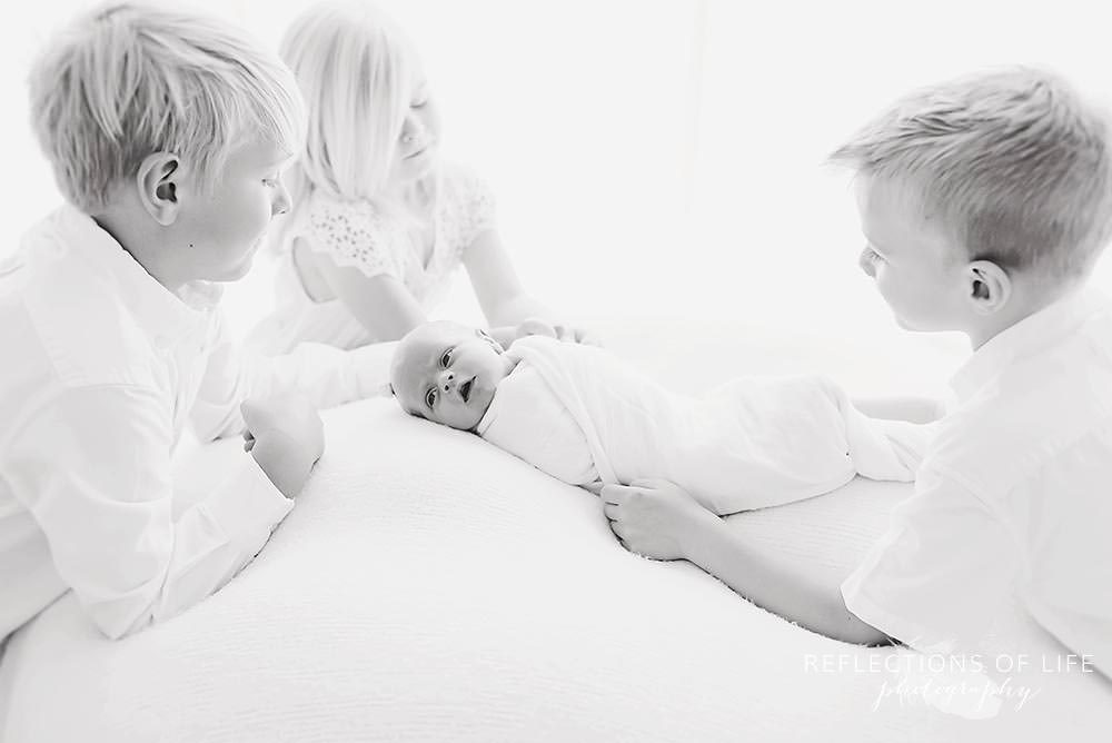 three siblings look at their new baby sister as she is wide awake in black and white