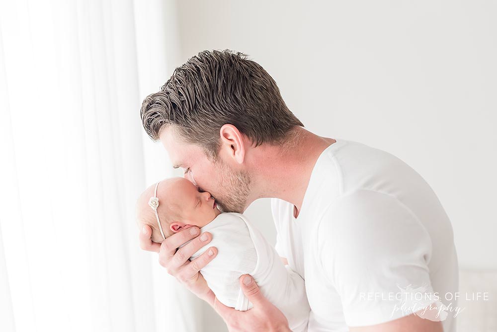 father holds his new baby close to his face and kisses her cheek