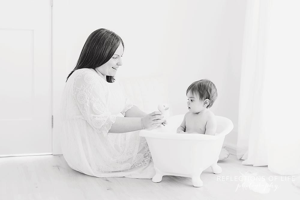 mama joins her baby in the bathtub in black and white and shows her a rubber duck