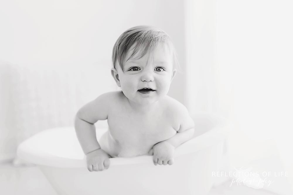 toddler smiles at the camera as he sits in the tub in black and white