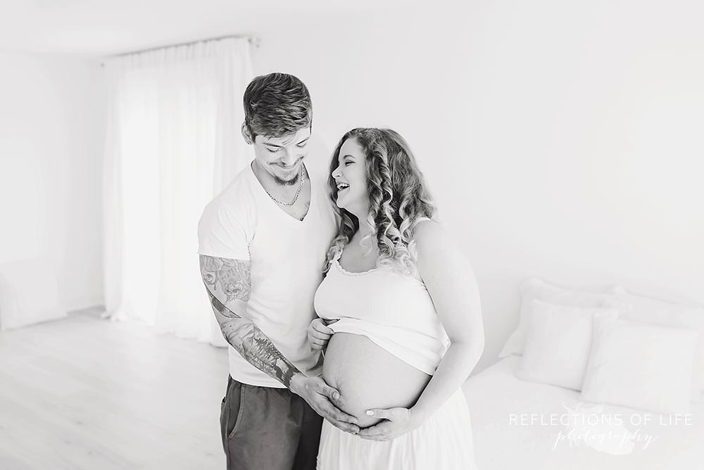 couple laughs together in natural light studio as dad holds her belly in black and white