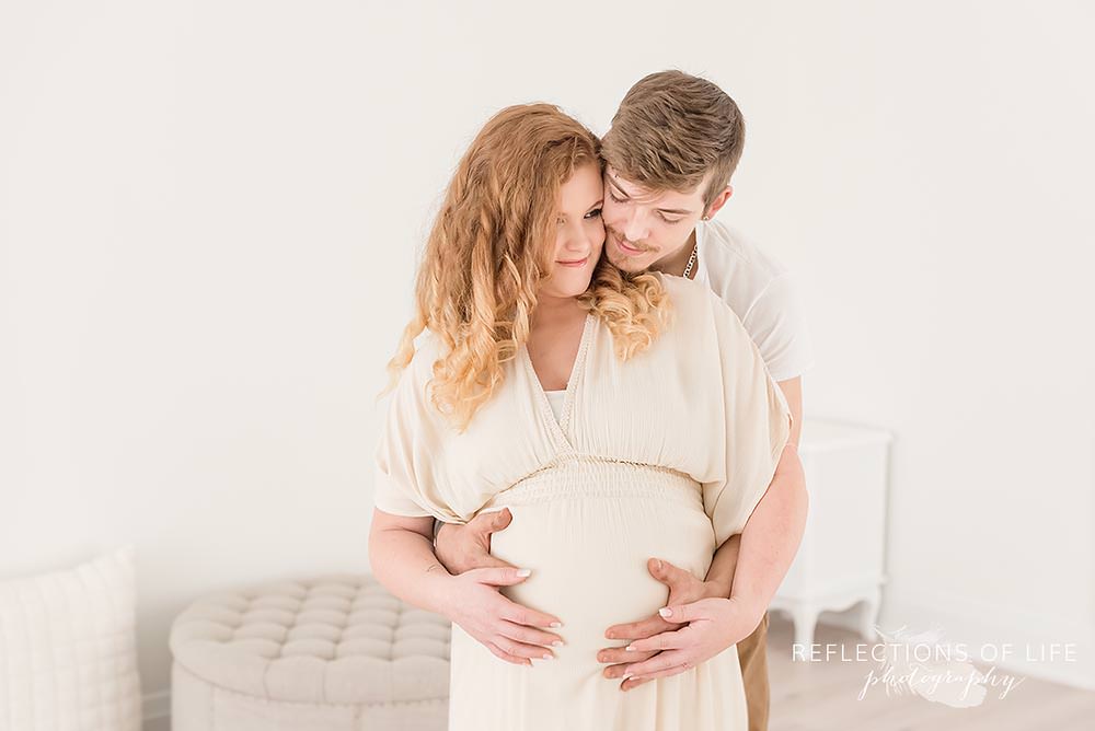 father holds mamas belly with her hands on his as they both smile in natural light studio