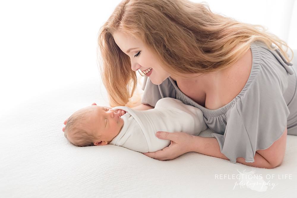 Baby swaddled in white blanket cuddling with beautiful mama in gray dress