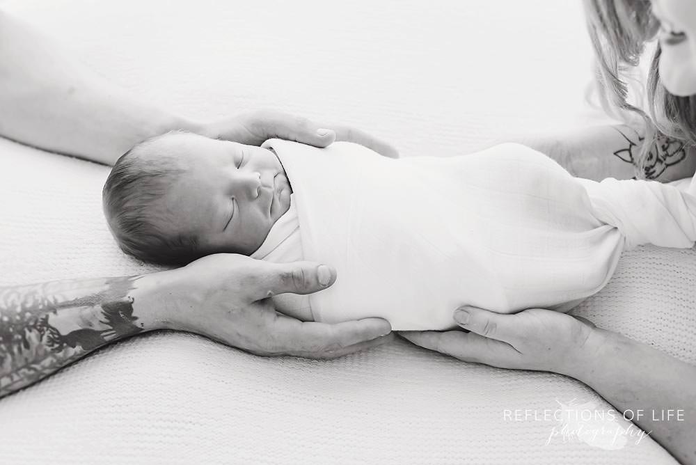 Baby swaddled in white with young parents hands laid on him