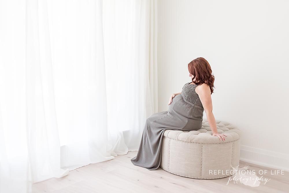 Expecting mama in gray dress sitting on linen ottoman near the window