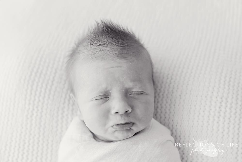 New born baby boy makes a sad face wrapped in white swaddle blanket natural light studio Grimsby