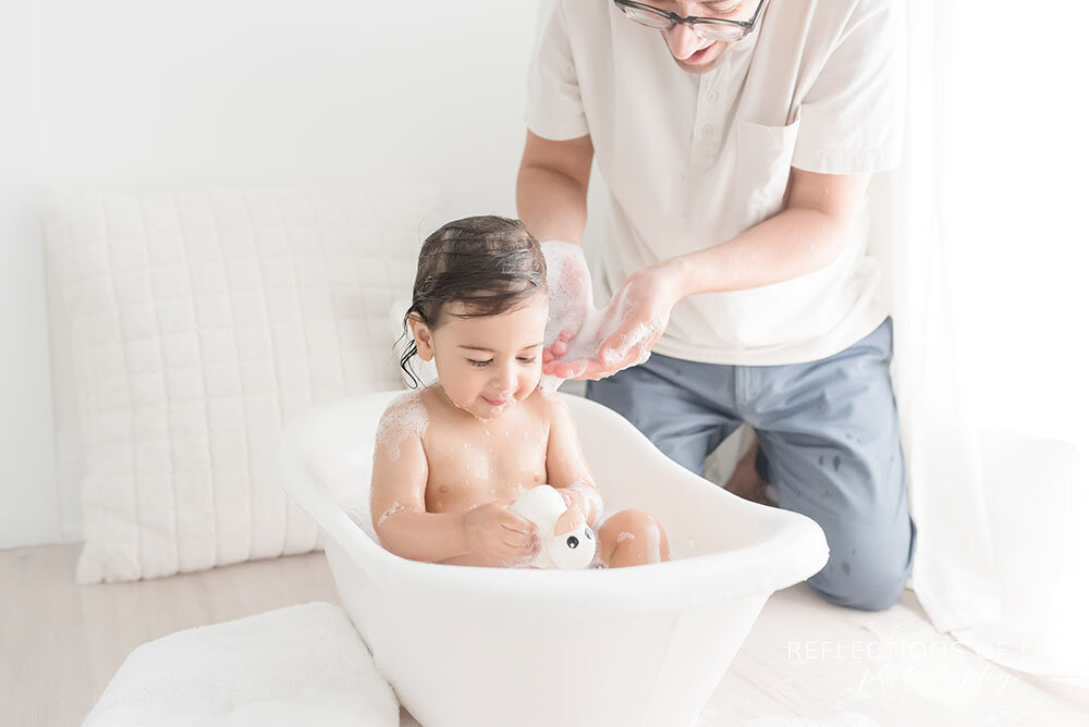 Daddy scoops warm water onto his sons shoulders in tiny bathtub in Niagara Region Ontario Canada
