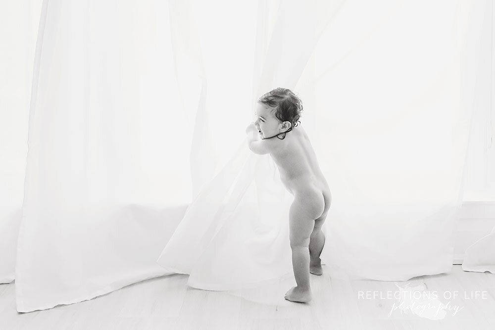 Naked baby boy playing in the curtains after photoshoot in natural light studio in Grimsby Ontario Canada