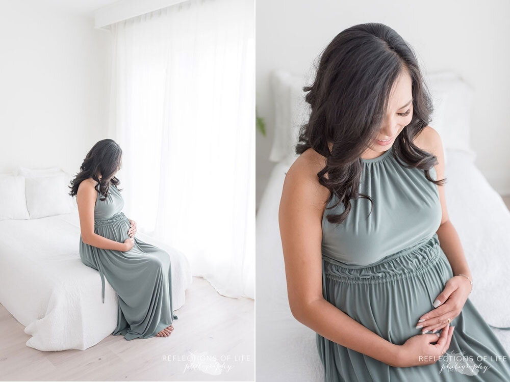 Green dress on gorgeous pregnant mama sitting on the edge of a bed with white comforter natural light