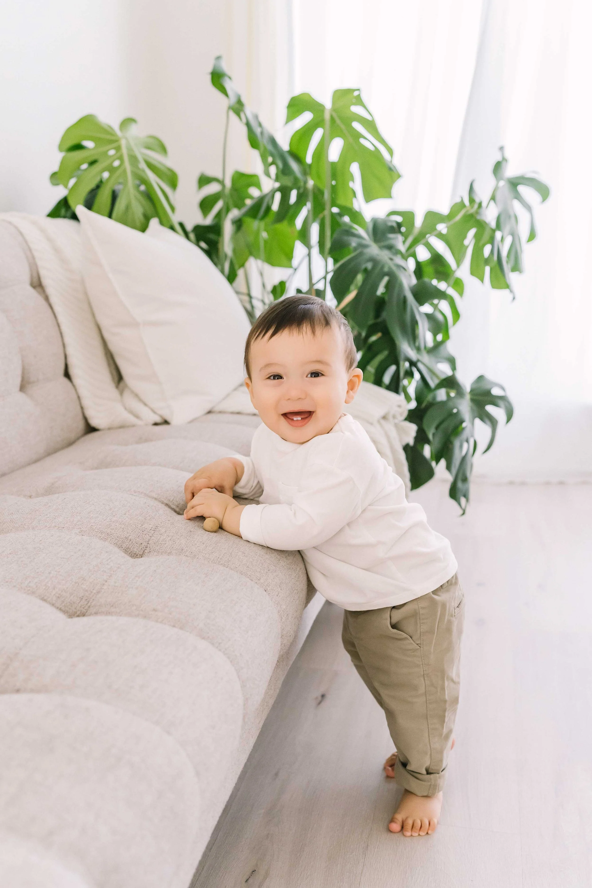 Energetic toddler smiling and pulling himself up on the couch during a relaxed first birthday photography session in Grimsby.