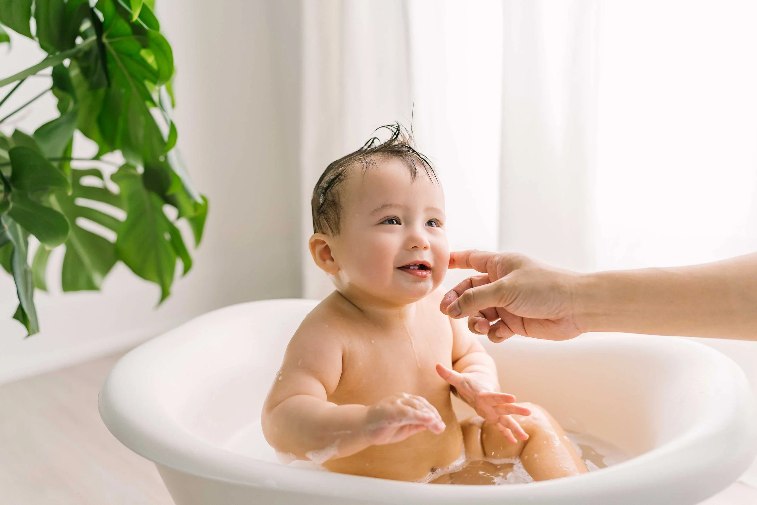 Playful baby photography image of a one-year-old boy laughing in a tiny bathtub during a fun bubble bath photoshoot after cake smash.