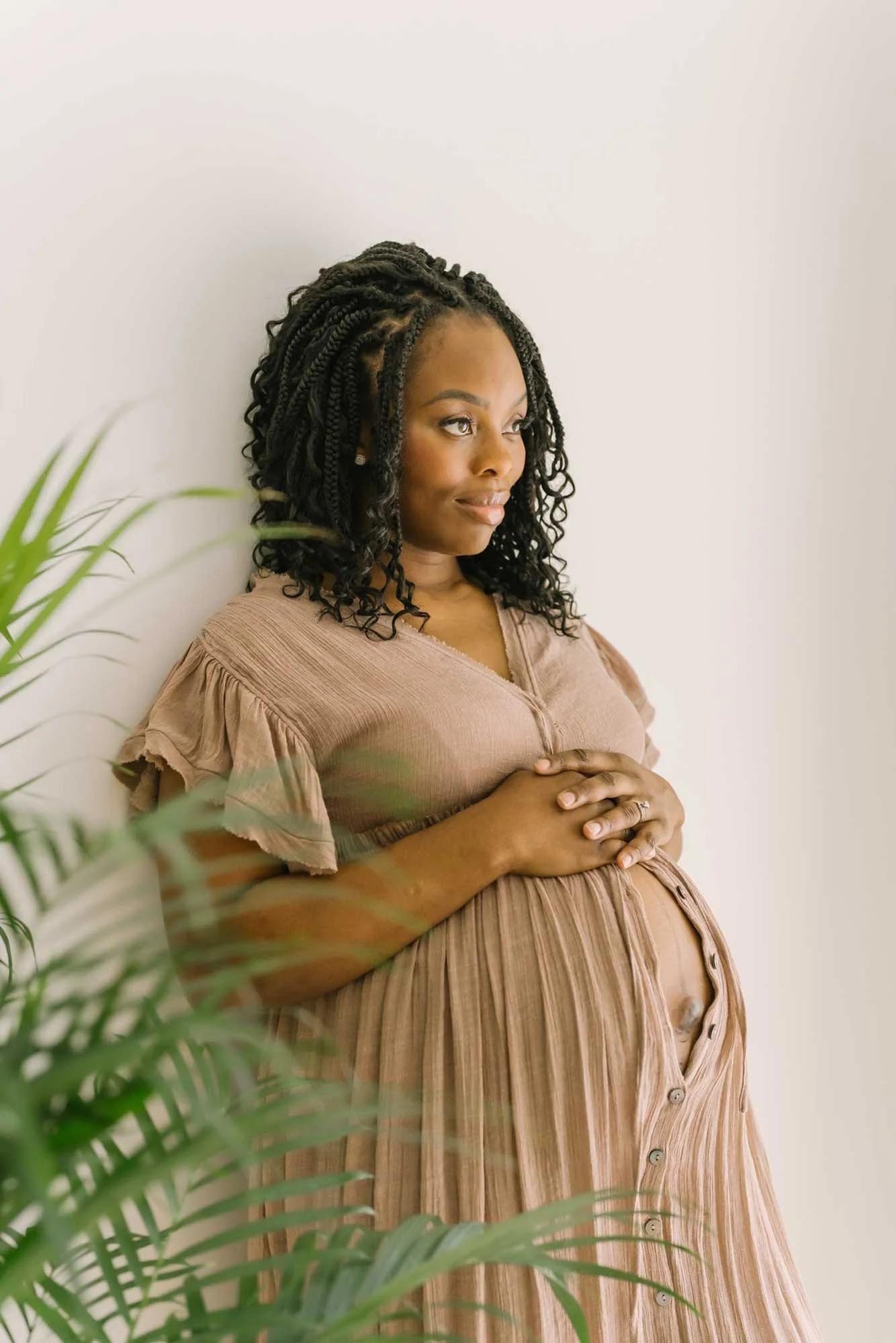 A serene studio maternity photo of a pregnant woman in a flowing dress, sitting on a beige armchair, gently cradling her belly with a peaceful expression taken by Karen Byker, newborn photographer in Niagara Region.