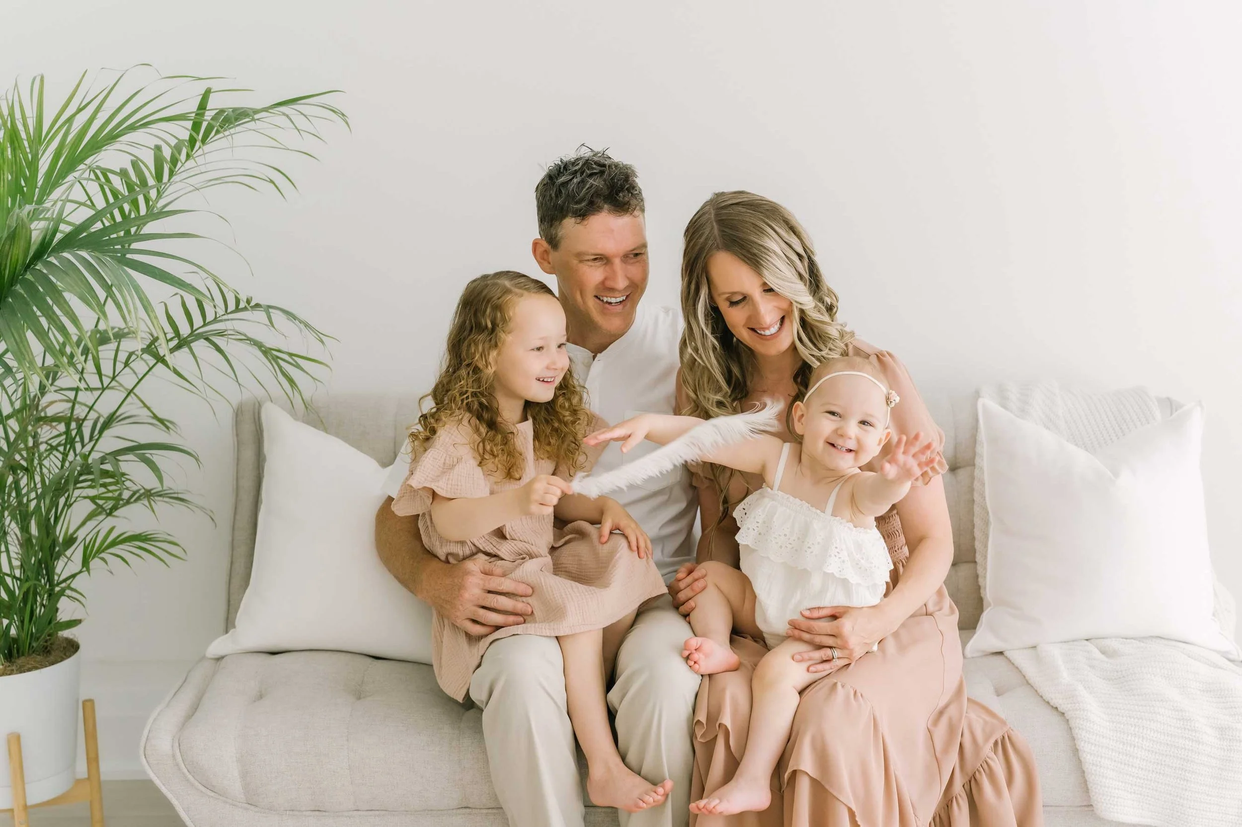 Fun family of 4 sits on couch in natural light studio tickling with a feather