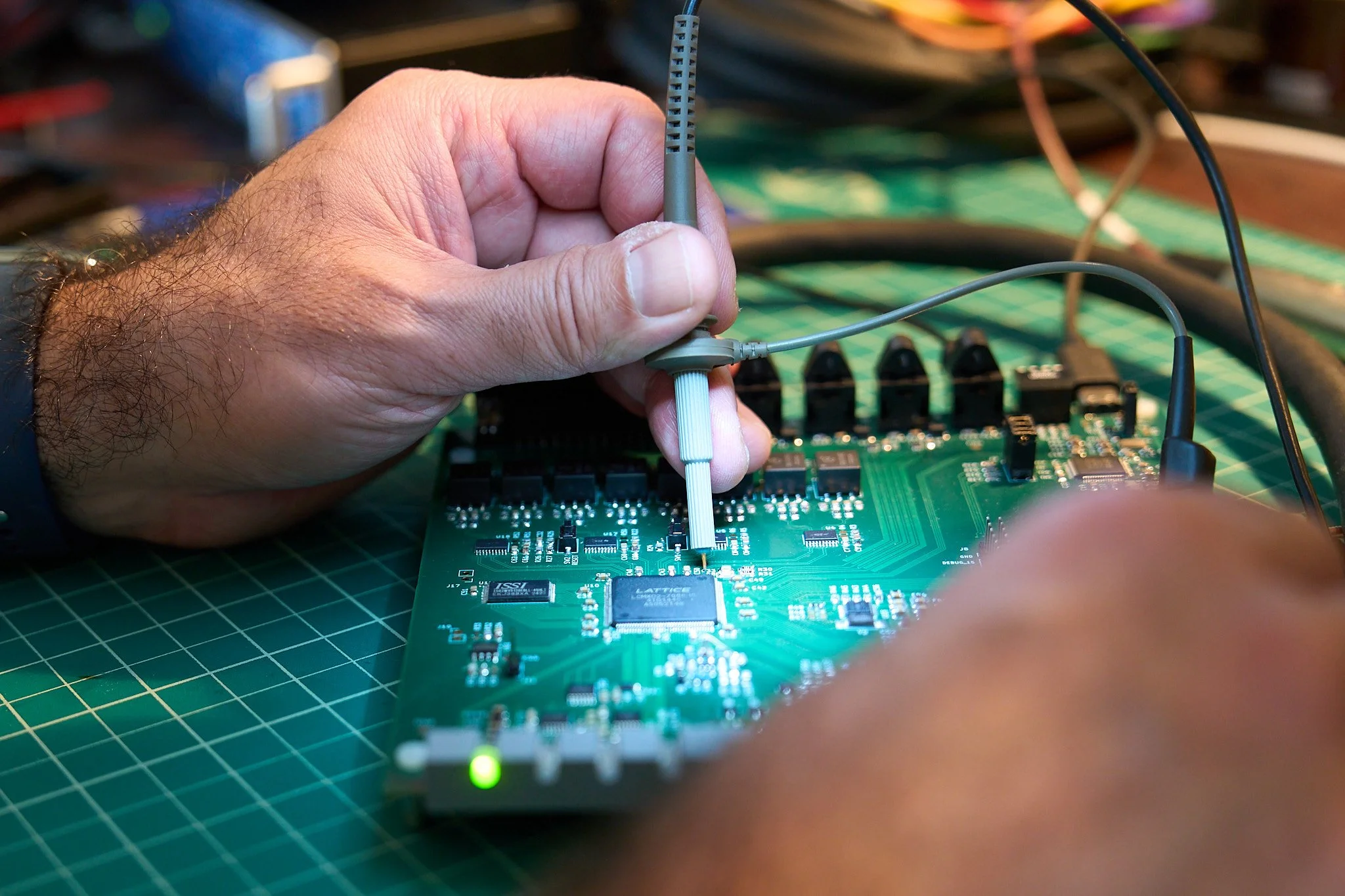 A person's hand is working on a green printed circuit board with electronic components, using a probe for assembly and testing. Cables are connected to the circuit board, and it is placed on a green ESD (electrostatic discharge) mat.