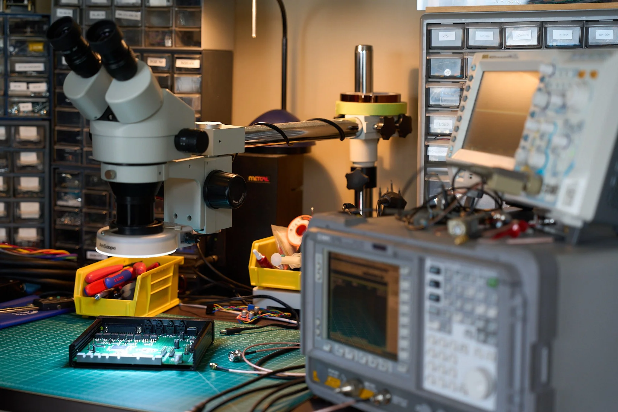 An electronics workbench with engineering equipment, including a microscope, oscilloscopes, and electronic components, surrounded by small plastic containers and tools.