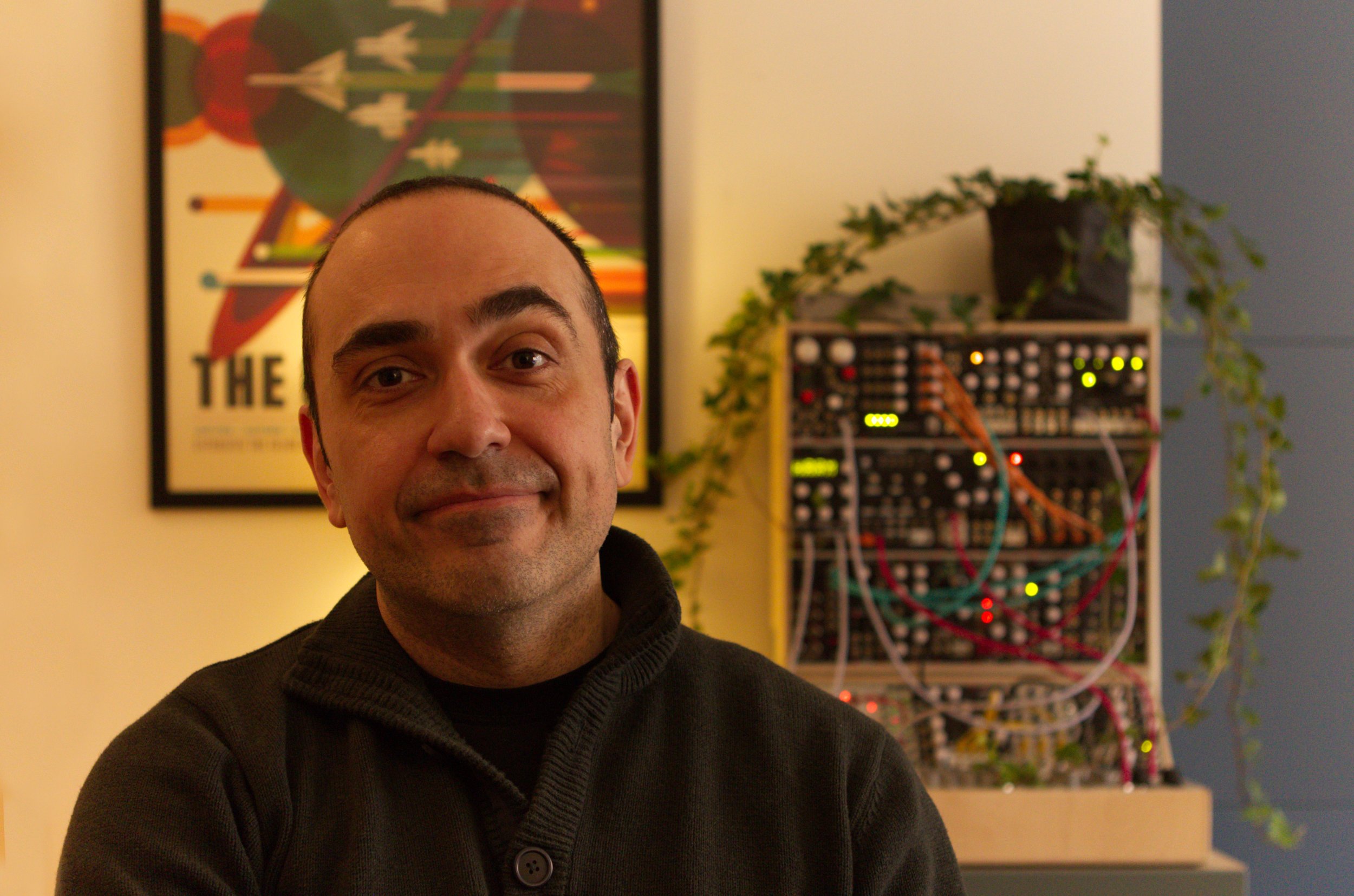 A man with short dark hair and a slight smile sitting in front of a Modular Synthesizer with colorful patch cables and lights in a room decorated with a framed poster and a houseplant.