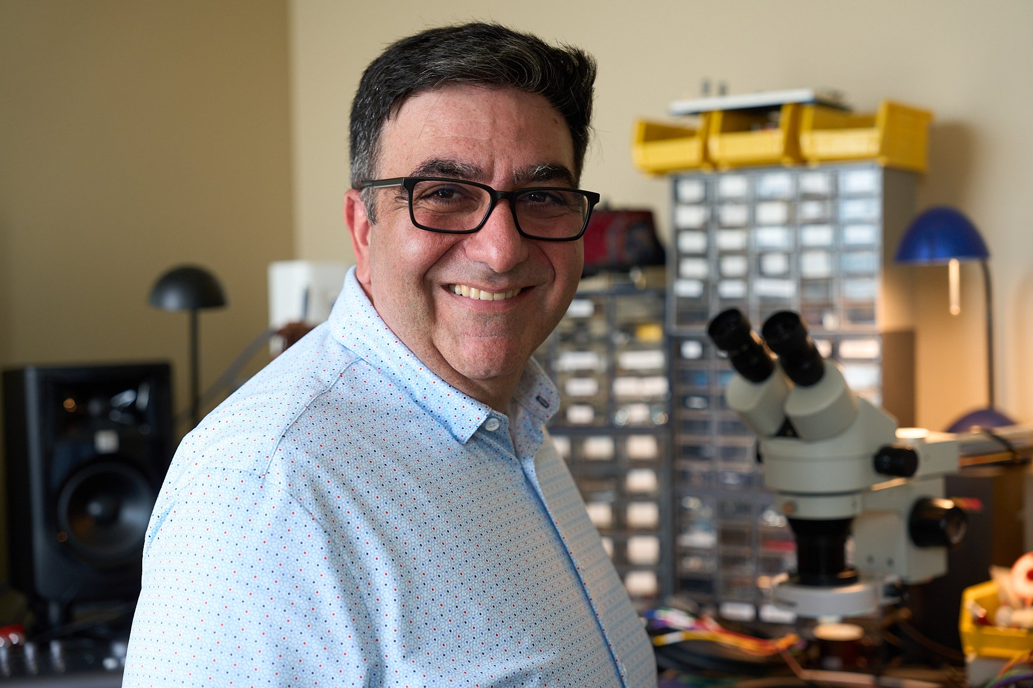 A man smiling, wearing glasses and a light blue shirt, in a workspace with electrical engineering equipment including a microscope, a speaker, and various storage containers.