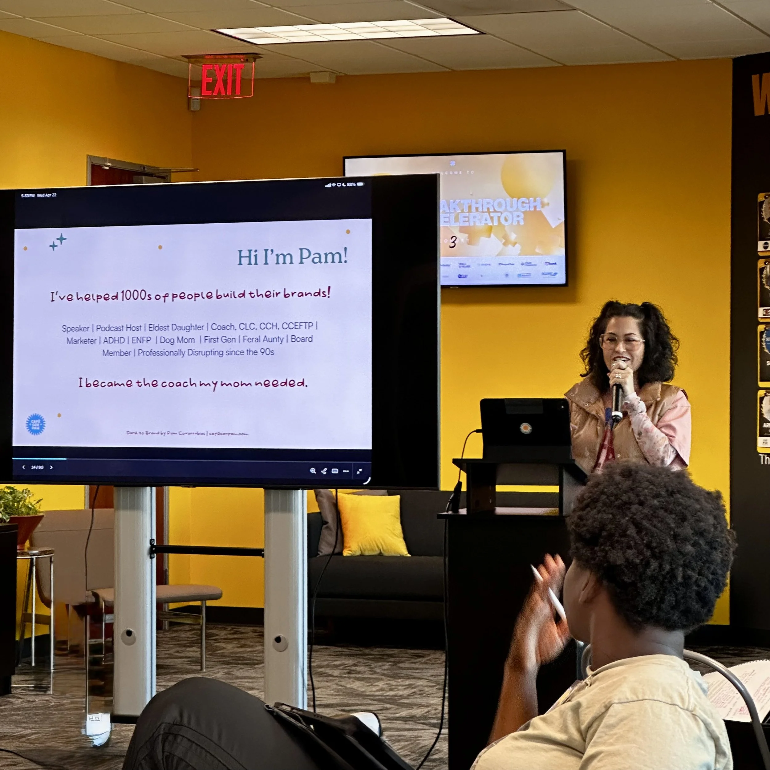 A woman in a pink floral shirt sitting at a conference table, speaking into a microphone. There is a large presentation screen behind her displaying a slide titled 'Hi I'm Pam!' with bullet points and colorful graphics. The setting appears to be a conference room with yellow walls, a ceiling light, and an emergency exit sign visible.