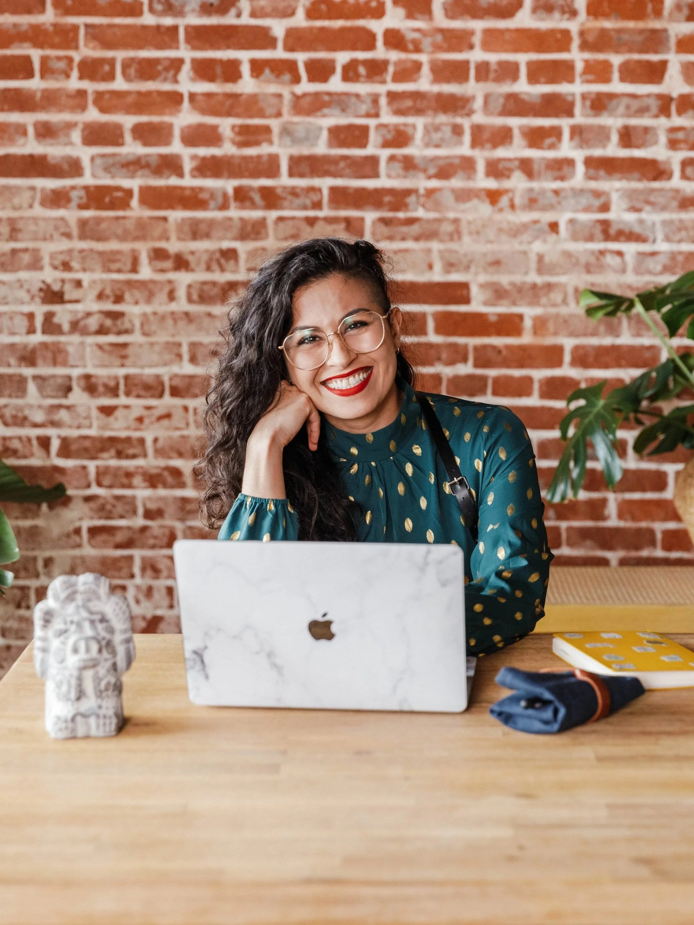 A woman with glasses, dark hair, and red lipstick smiling and resting her chin on her hand, sitting at a wooden table with a marble laptop in front of her, and a brick wall in the background.