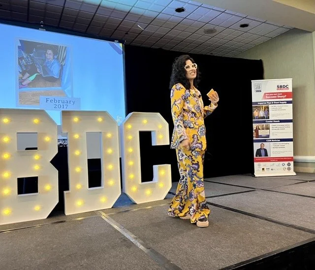 A woman standing on a stage in front of large illuminated letters spelling 'SEO.' She is holding a microphone and a note card, wearing a colorful patterned outfit. Behind her is a screen displaying February 2017, and a poster with information about an SBDC event.