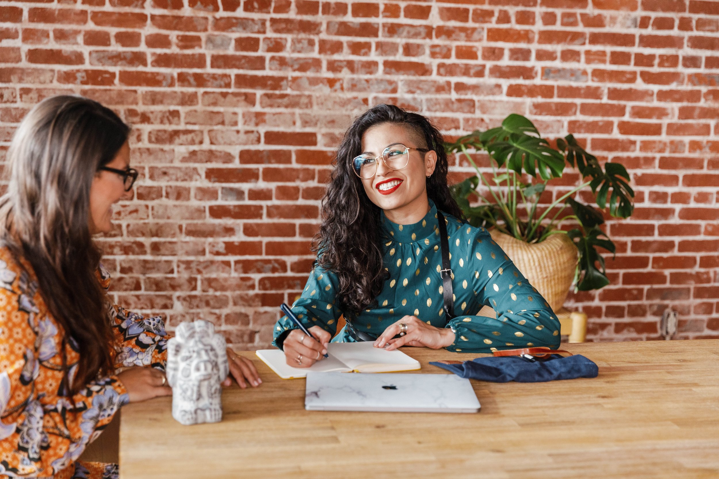 Two women sitting at a wooden table in a room with a brick wall. One woman has long dark hair, glasses, and a patterned blouse. The other woman has curly black hair, glasses, and a teal blouse with gold dots. The woman with curly hair is smiling and writing in a notebook. There is a potted plant with large green leaves in the background.