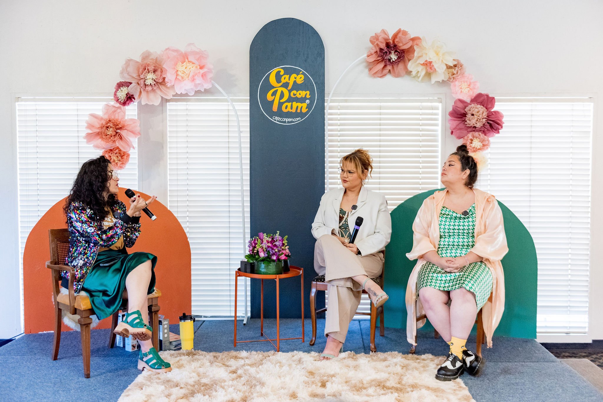 Four women sitting on colorful chairs in a room decorated with flower-themed balloons and a sign that reads 'Café Con Pam.' One woman is speaking into a microphone, while the other three women listen and engage in conversation. There is a small table with flowers and a purple bag, and a soft cream carpet on the floor.