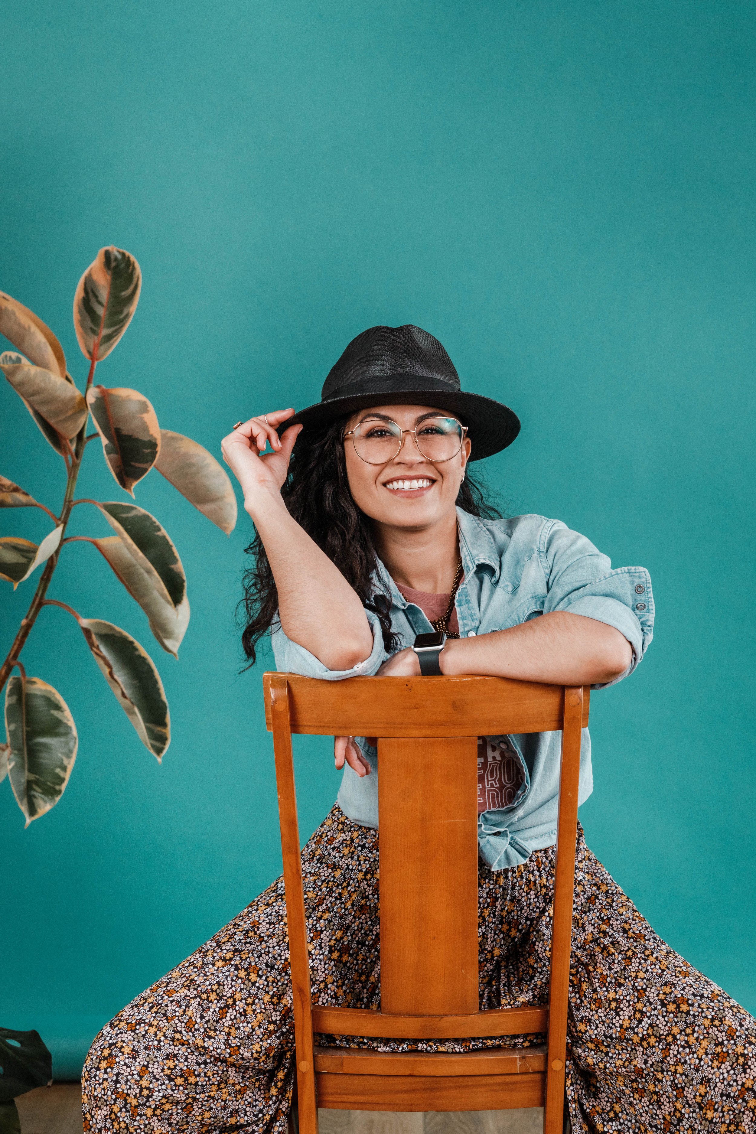 A smiling woman wearing glasses, a black hat, and a denim jacket, sitting on a wooden chair with the backrest visible, against a teal background. A leafy plant is on the left side of the image.