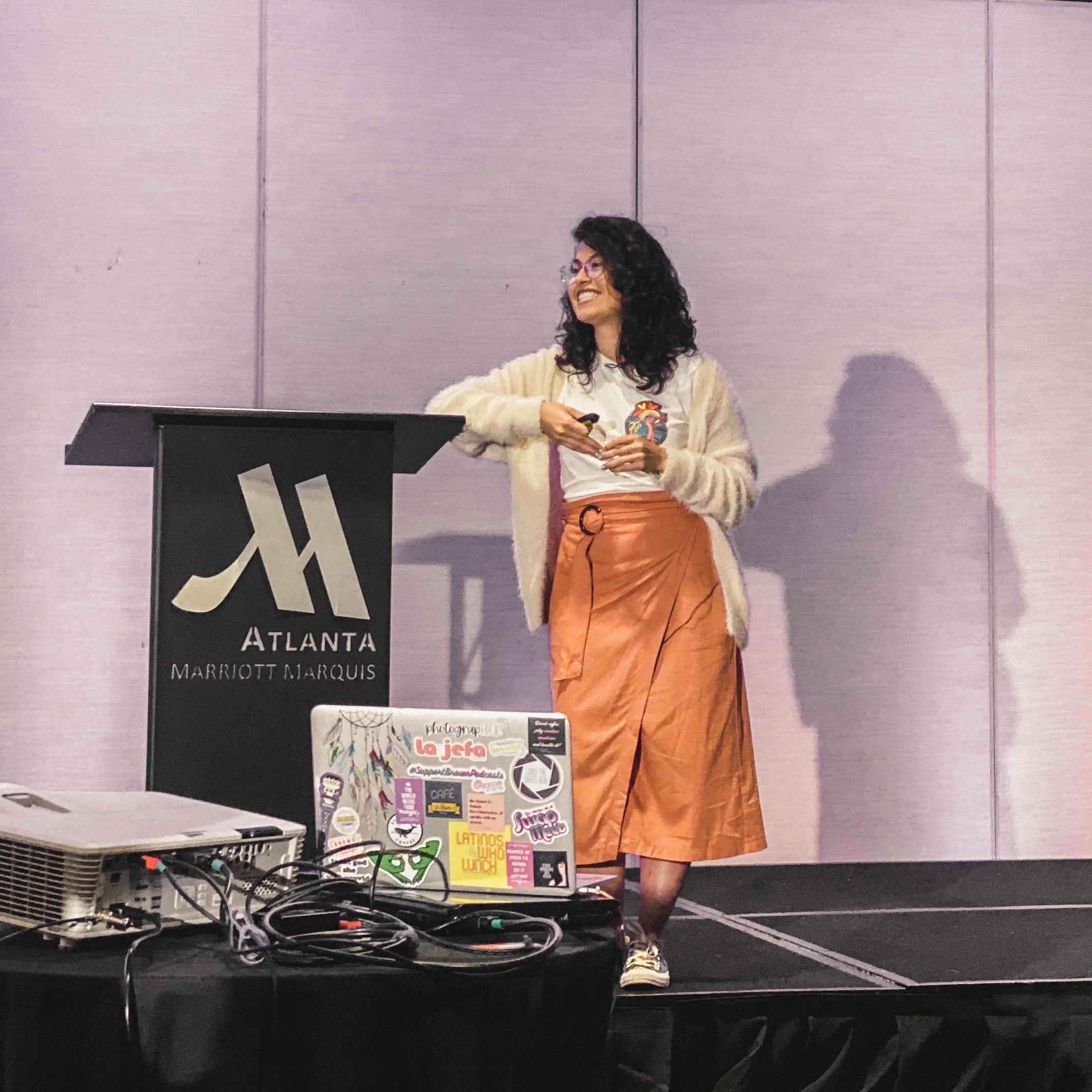 A woman with curly dark hair, glasses, and a big smile, standing behind a DJ setup with various stickers on a laptop, in front of a lavender-colored wall at an event or conference.