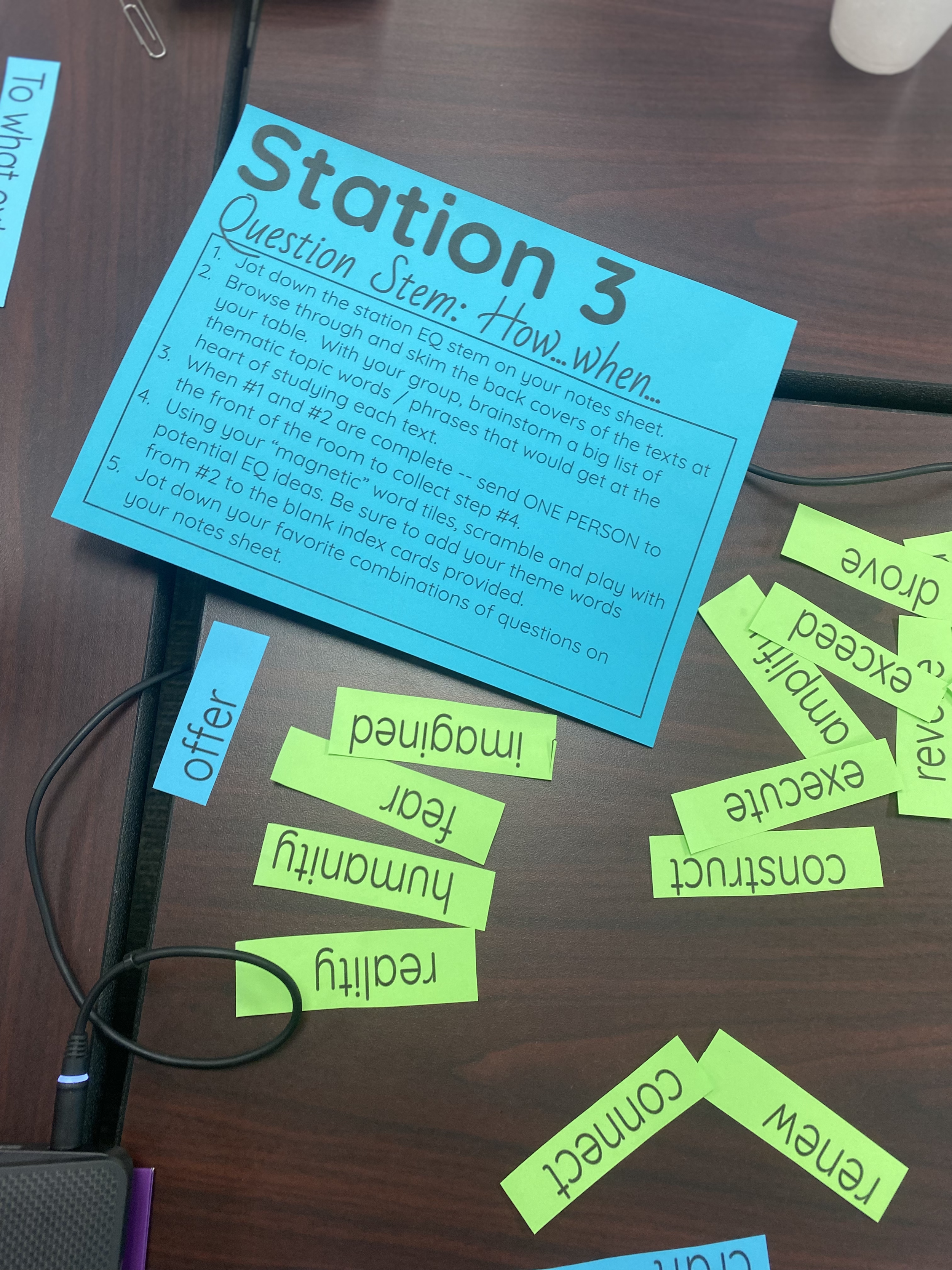 A blue sheet of paper titled 'Station 3 Question Stem: How ... when...' with numbered instructions. Surrounding it are green and blue slips of paper with words like 'review,' 'exchange,' 'humility,' 'teach,' 'confusion,' and 'awake.' A purple pencil 