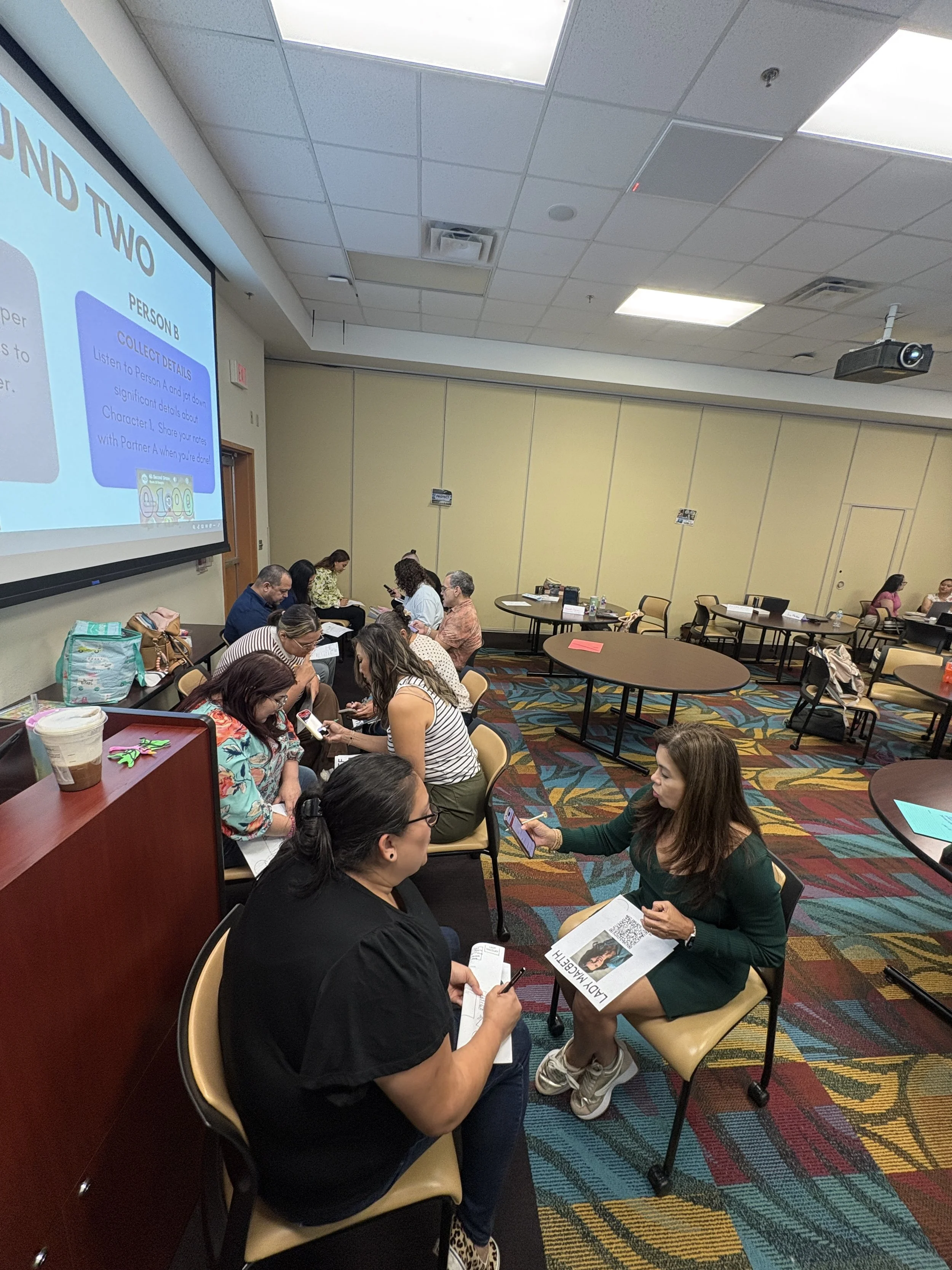 People sitting and talking in a conference room, some examining papers or using their smartphones, with a large projection screen displaying a presentation on the left.