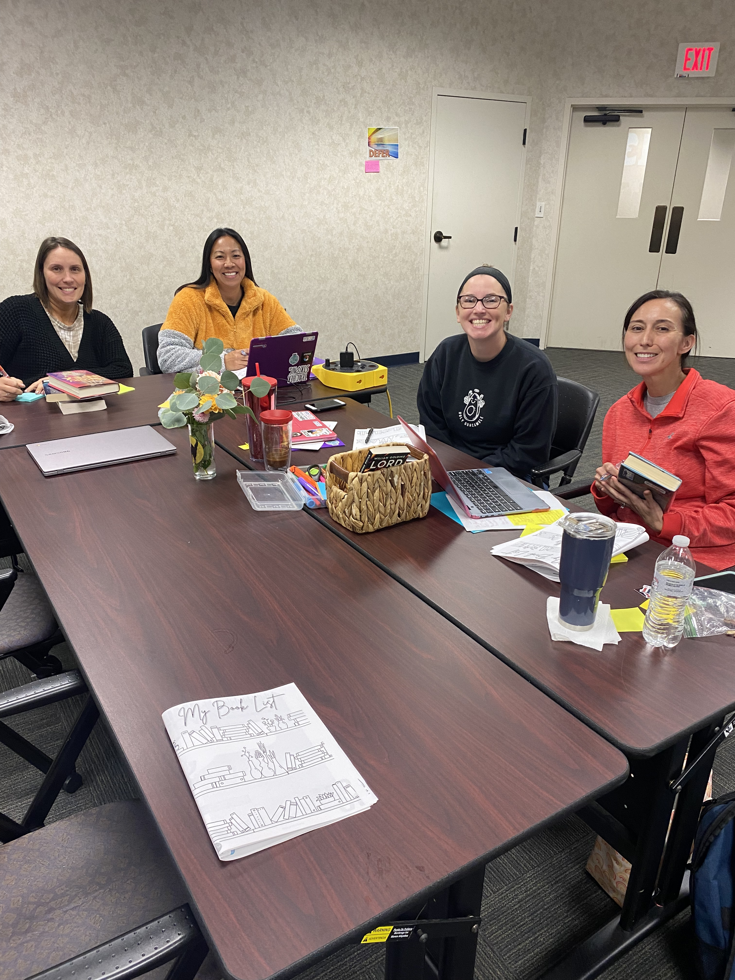 A group of four women sitting around a rectangular conference table in a meeting room, smiling at the camera. The table has notebooks, papers, a laptop, a basket, pens, a water bottle, and a small plant.