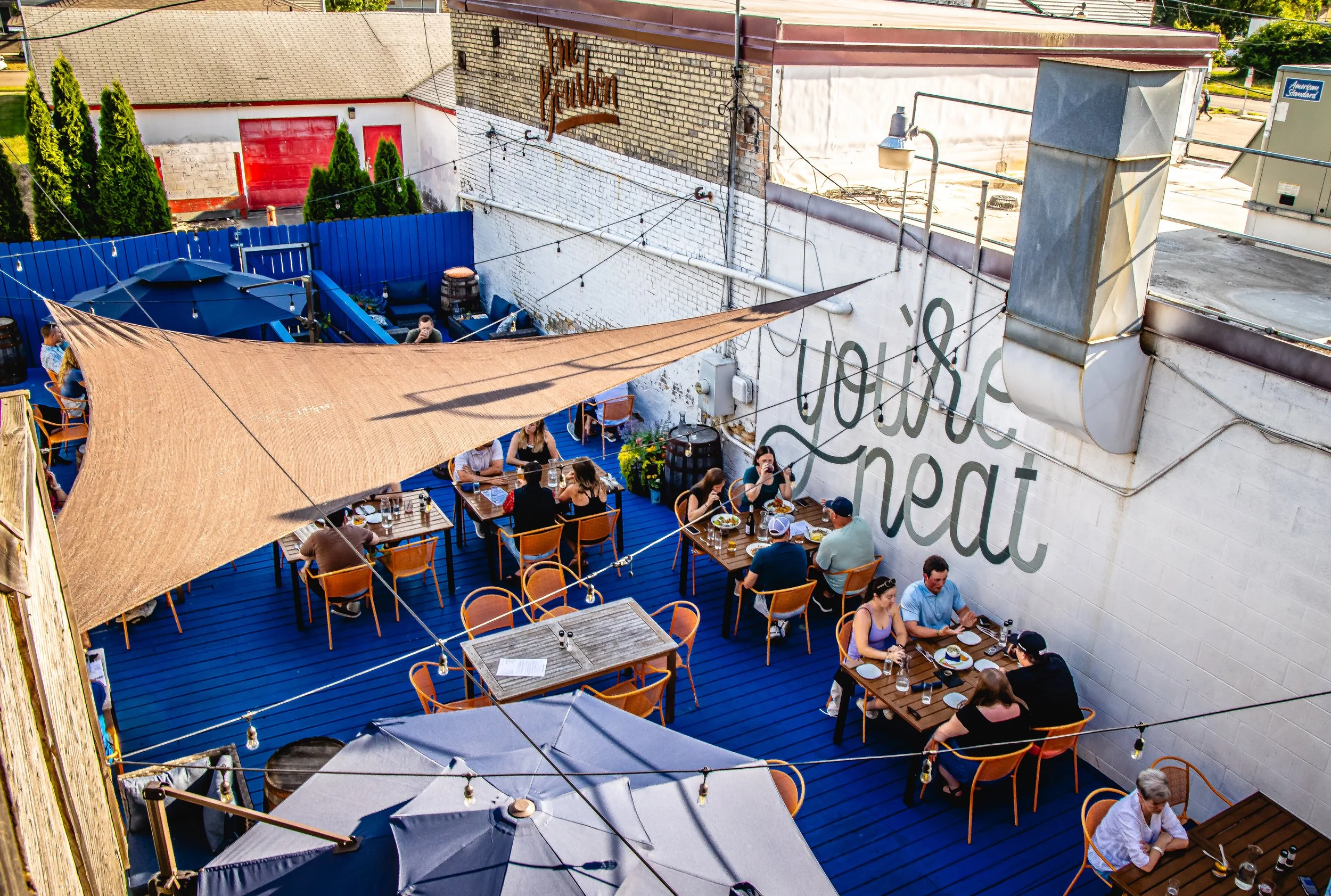 Outdoor rooftop dining area with tables, chairs, umbrellas, and string lights, surrounded by brick and painted walls, with people eating and socializing.