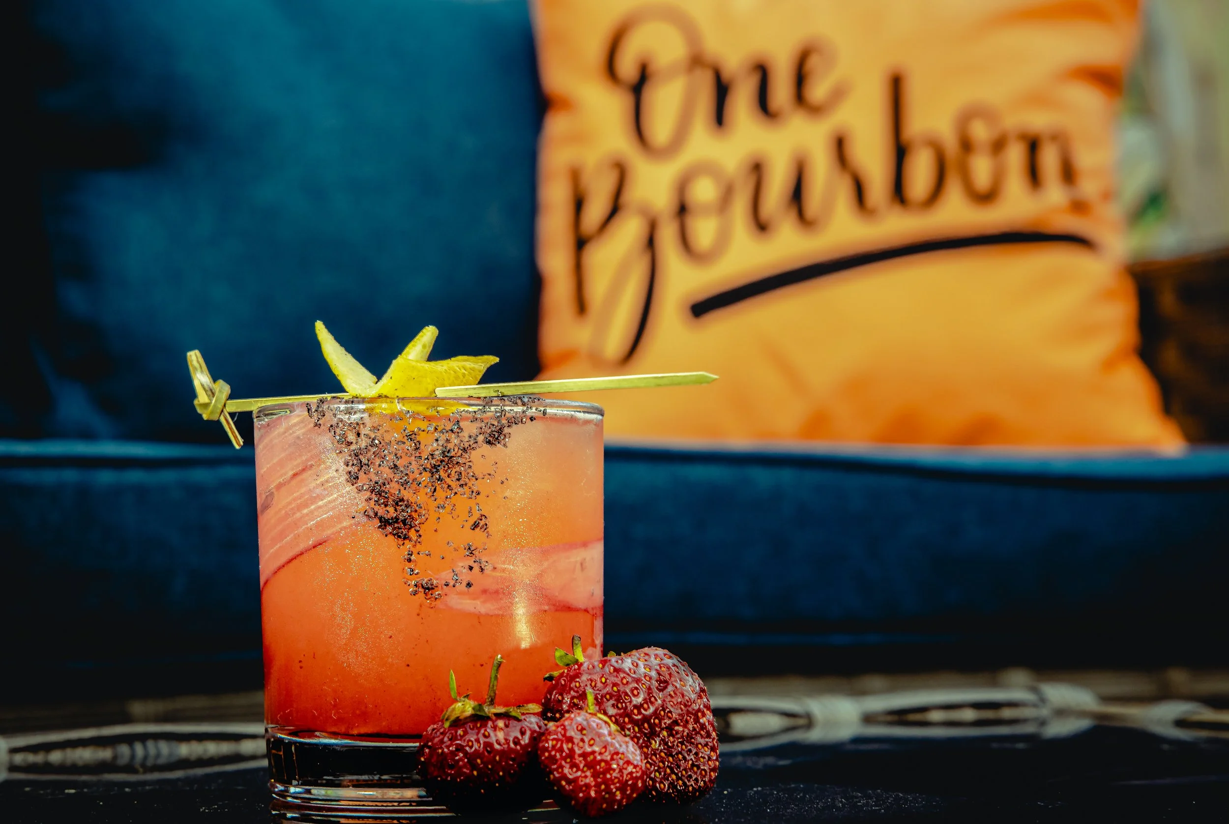 A pink cocktail in a glass garnished with a lemon twist and sprinkled with black salt, with fresh strawberries around the base of the glass, and a blurry orange sign in the background that reads 'The Feather.'