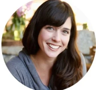 Smiling woman with long dark hair wearing a gray top, outdoors with blurred background of greenery and sunlight.