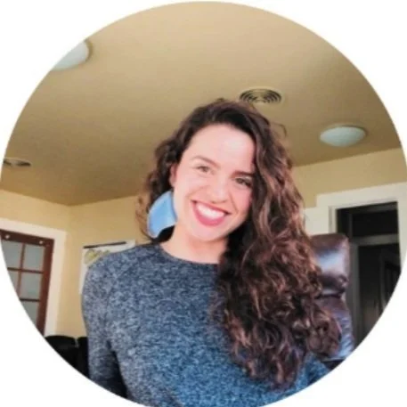 Smiling person with curly hair wearing a gray sweater, sitting indoors in an office-like room.