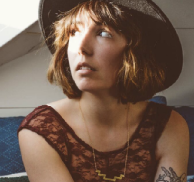 Woman with short brown hair wearing a black hat and a lace top, gazing to the side, sitting indoors in natural light.