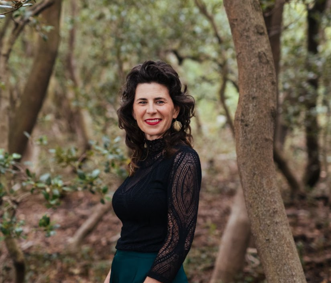 Woman in black lace top standing outdoors in a wooded area.