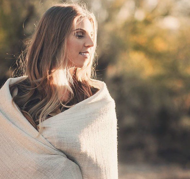 Woman wrapped in a light-colored blanket outdoors, surrounded by blurred nature background.